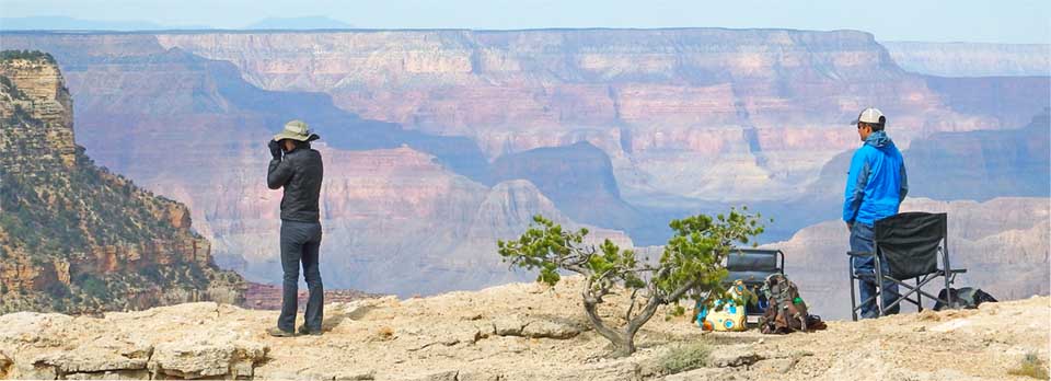 A man and a woman with binoculars standing on a rock ledge overlooking Grand Canyon. They are looking for raptors. A man and a woman with binoculars standing on a rock ledge overlooking Grand Canyon. They are looking for raptors.