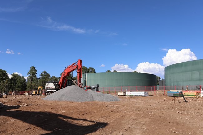 A view of construction equipment preparing a dirt construction site for the concrete foundation. Two water tank structures are in the background.