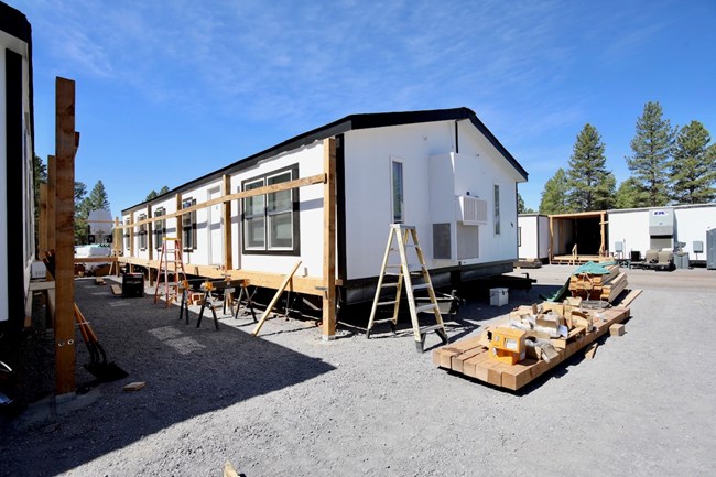 Wood framing is being installed to create covered decks between two trailers. Piles of wood and other construction materials are visible in the foreground.