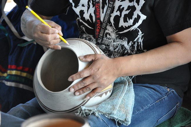 American Indian artist holds pale bowl in lap. One hand holds the bowl while the other hand traces designs and patterns onto the bowl.