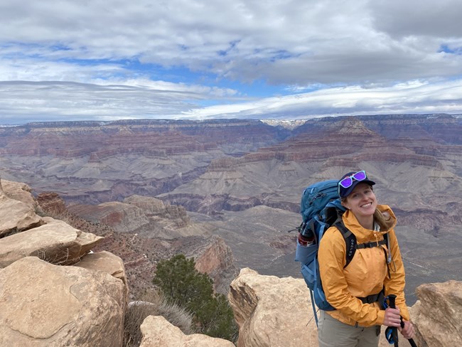A woman with a yellow jacket and blue backpacking pack smiles in front of colorful canyon walls and cliffs of the Grand Canyon.