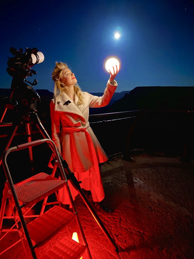 A woman holds an illuminated ball next to the full moon at dusk