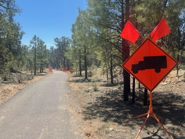 Greenway Trail truck crossing signage An orange construction sign with a large black truck is placed alongside a trail to indicate a construction zone and crossing is ahead