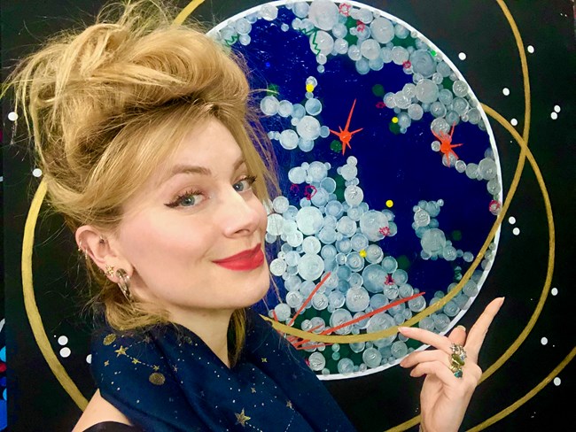 A woman with blonde hair and red lipstick points to a artistic display of the solar system behind her