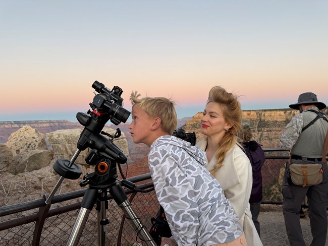 A woman looks at a child as he looks through a telescope at dusk