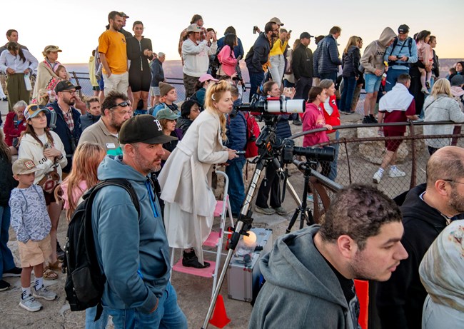 A crowd outside looks on together at a rising moon. An astronomer stands poised with a telescope in the middle of the crowd.