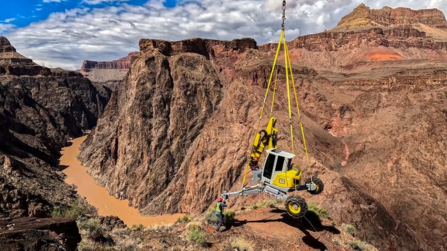 A helicopter is transporting an excavator in Grand Canyon National Park. Canyon walls and the Colorado river are visible in the background.