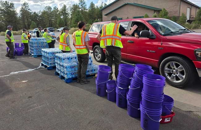 Workers in yellow vests assist in distributing pallets of water, buckets, and dishpans at a water distribution event