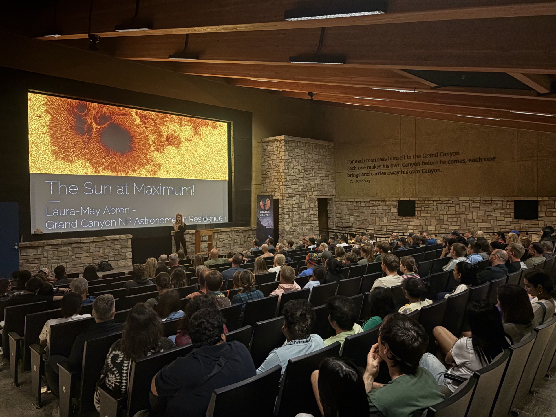 A packed indoor theater with a woman speaking at the front