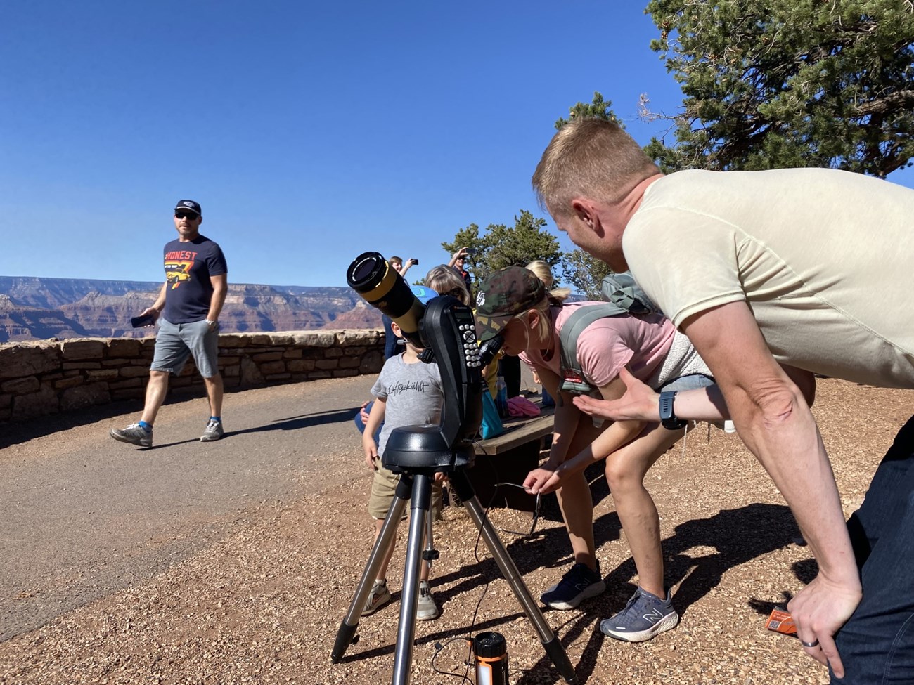 A man with red hair and a yellow shirt stands over a small telescope during the day while a family looks through it.