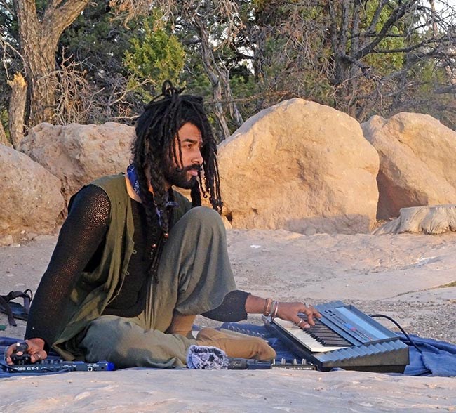 a musician with long black hair in dreadlocks is playing a keyboard outside with large boulders and trees in the background.