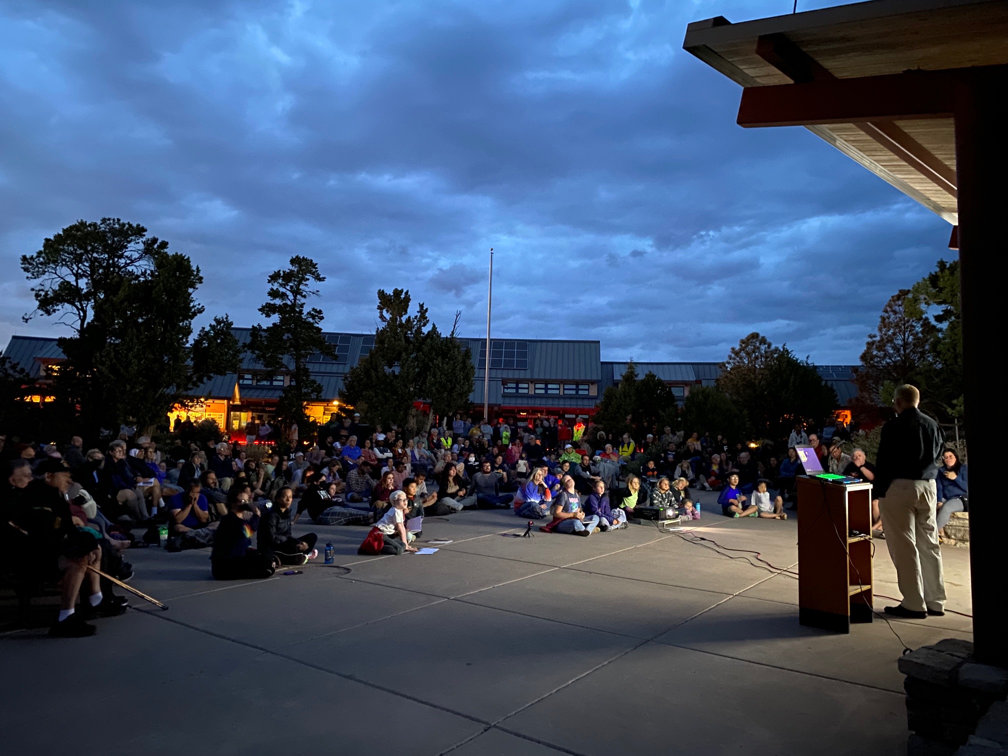 A crowd sits on pavement outside. A man stands in front of a projector.