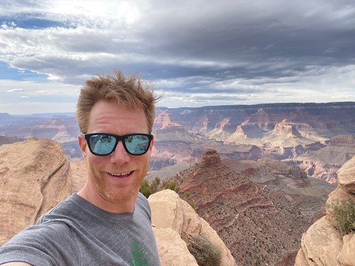 A man with sunglasses and red hair takes a selfie in front of colorful canyon walls and cliffs.