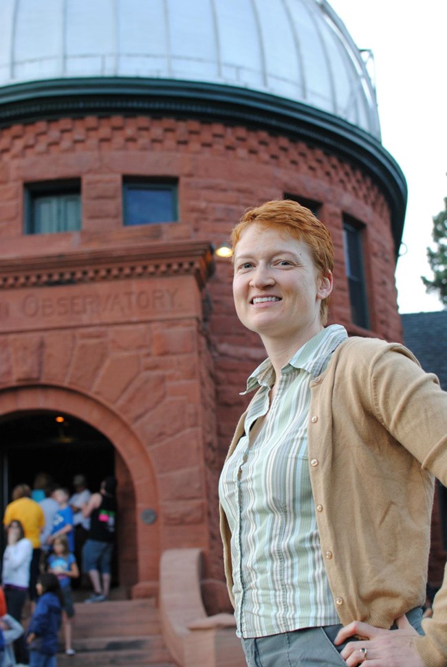 A woman with short red hair stands in front of a stone observatory.