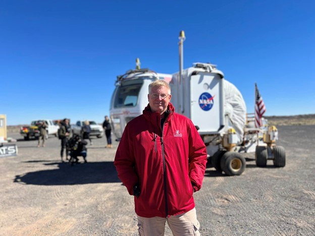 A man in a red jacket stands outside in front of a white NASA roving vehicle.