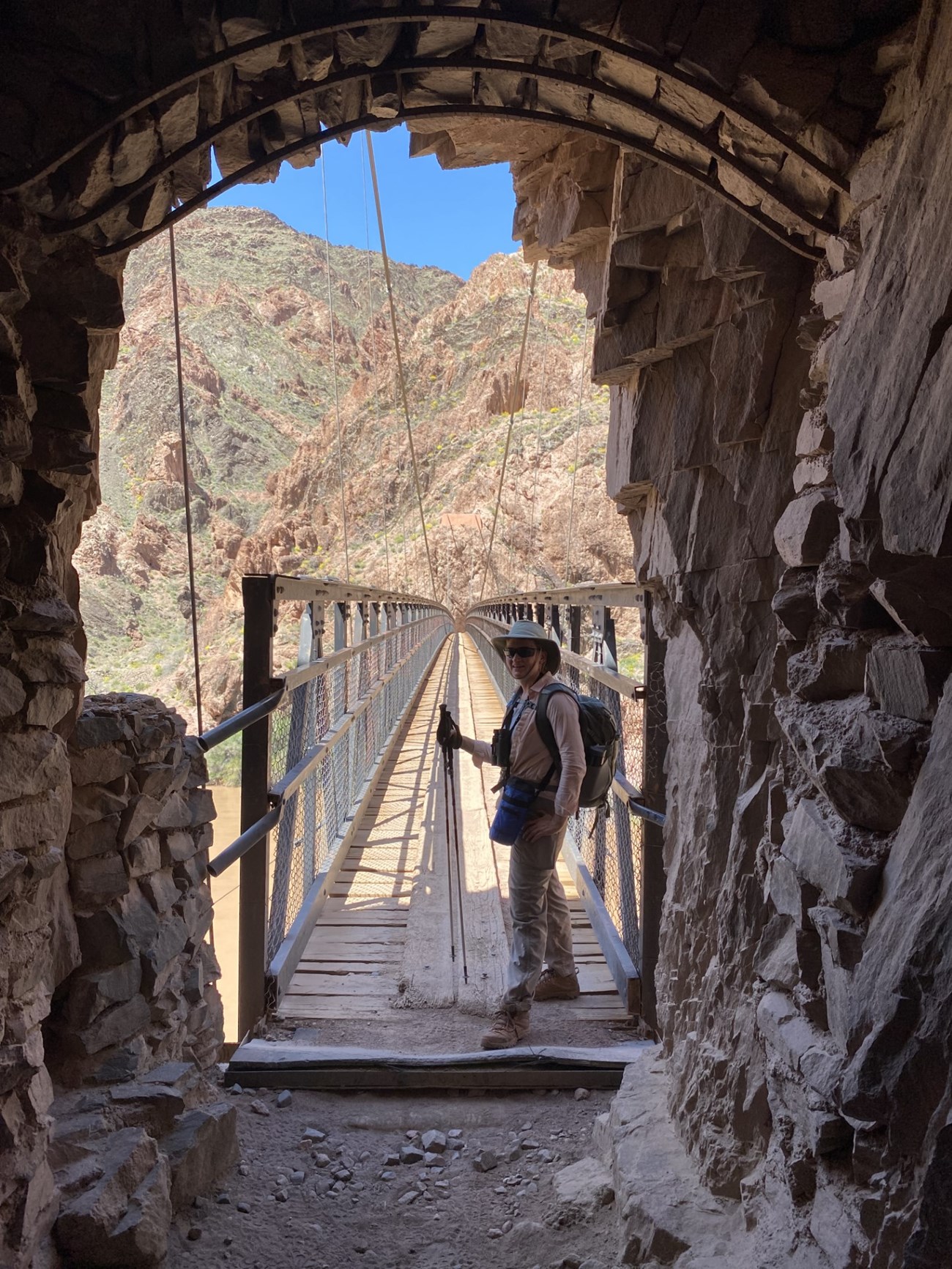 A woman in hiking gear stands in front of a rock opening out to a black suspension bridge.