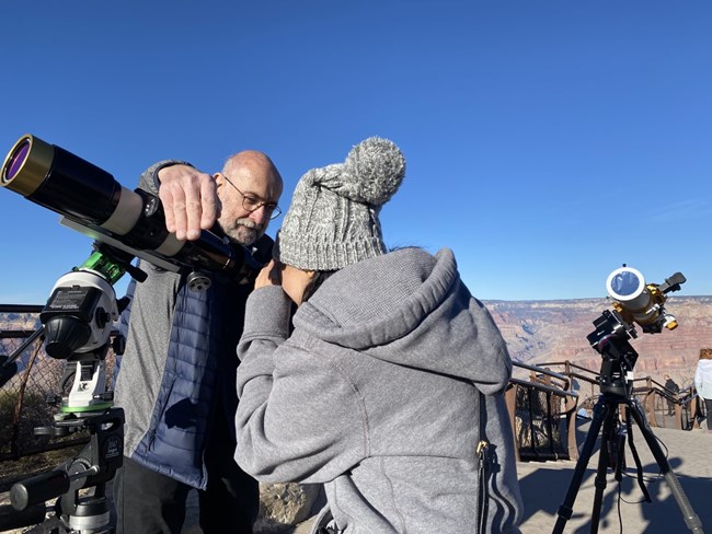 A man stands next to a telescope. A woman with a grey coat looks into the solar telescope. Colorful canyon walls in the backgound.