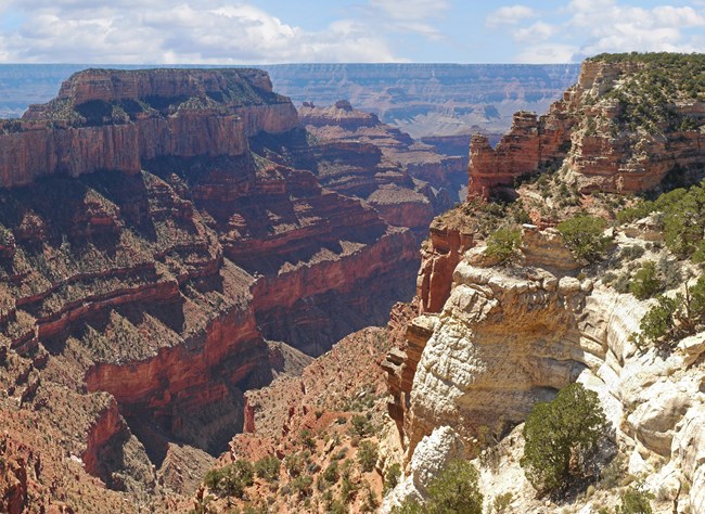 a sandy-colored rock column juts out against a distant series of stratified cliffs rising to a flat-top mountain.