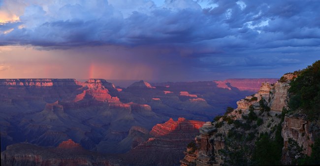 The Grand Canyon, illuminated by red light and purple shadows with a layer of dark blue clouds hanging overhead. The trace of a partial rainbow.