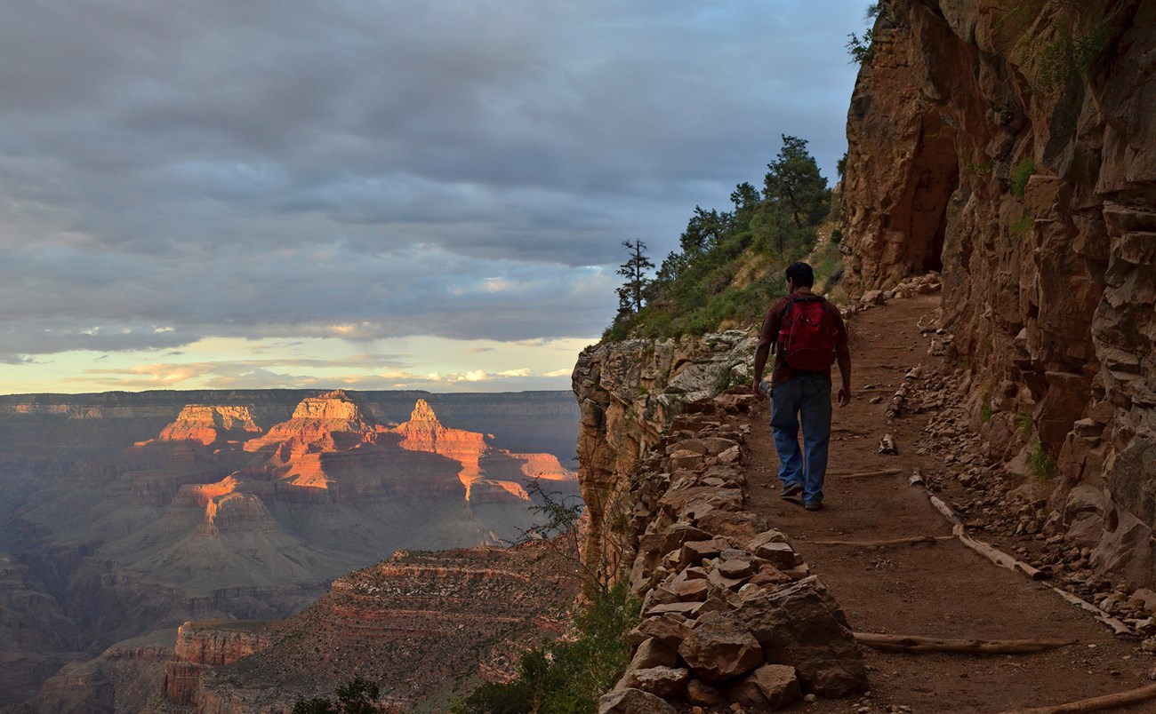 A single man with a backpack hikes uphill, towards a golden sunset background.
