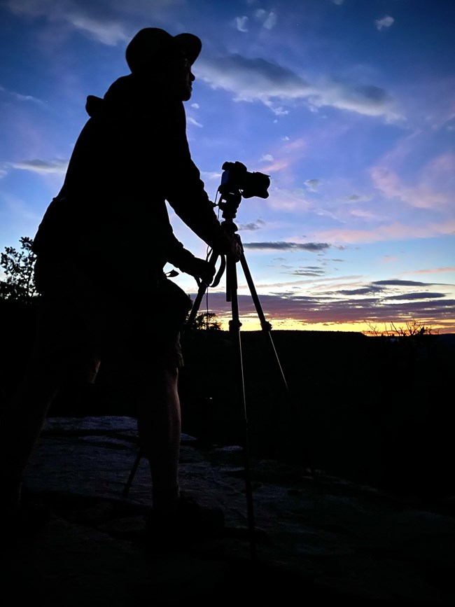 A silhouette of a man with a camera on a tripod. A colorful canyon sunset sky in the background.