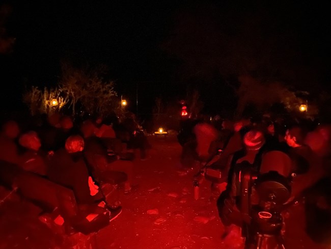 An audience sits at night shrouded in red light. The aisle is in the center of the image. A man stands at the end of the aisle with a red light.