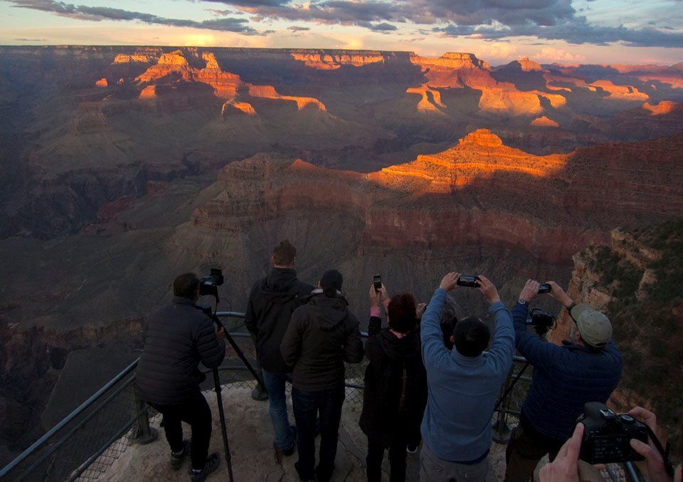 A group of visitors stand facing a sunset-lit canyon, photographing it with smart phones and traditional cameras.