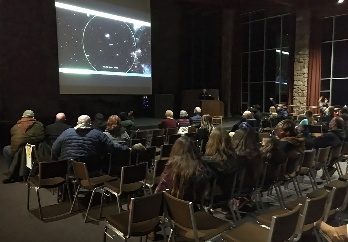 An audience sits in an auditorium. An image of the solar system is displayed on a large screen at the front.