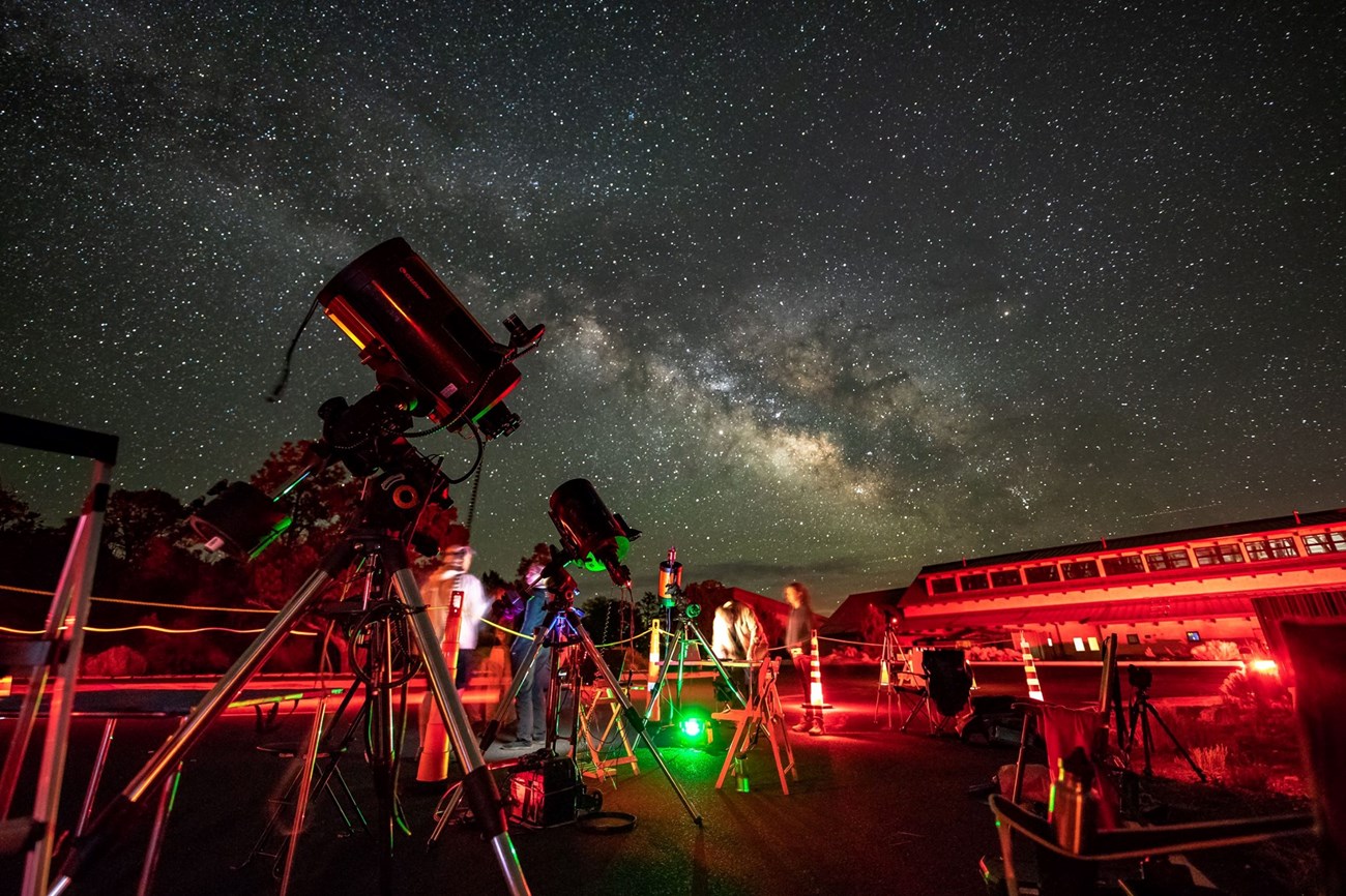 Some telescopes appear in the foreground. The Milky Way is in the background. People stand around the scopes.