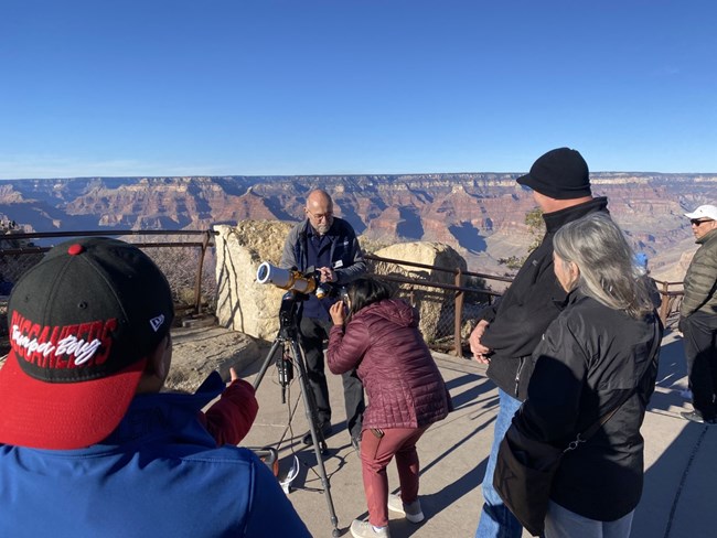 A man stands next to a solar telescope on the rim of the Grand Canyon. Several visitors gather around.