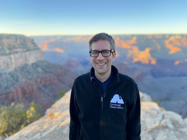A man stands smiling with a black sweater that says "Cincinnati Observatory" on it. The background shows out of focus colorful canyon walls and cliffs of red and white.