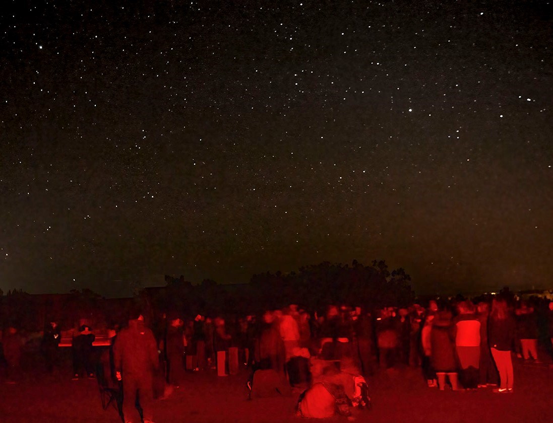 A red-lit audience stands outside under a sky full of stars.