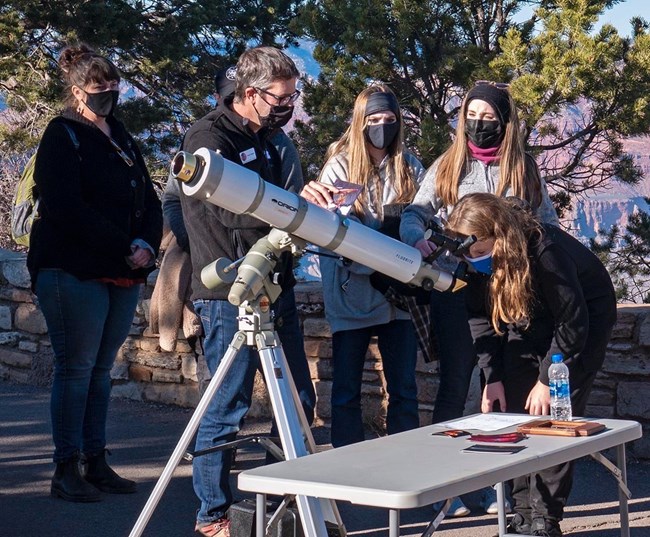 A man stands next to a long white telescope in the daytime. Three masked women stand in a line, one woman looks through the scope.