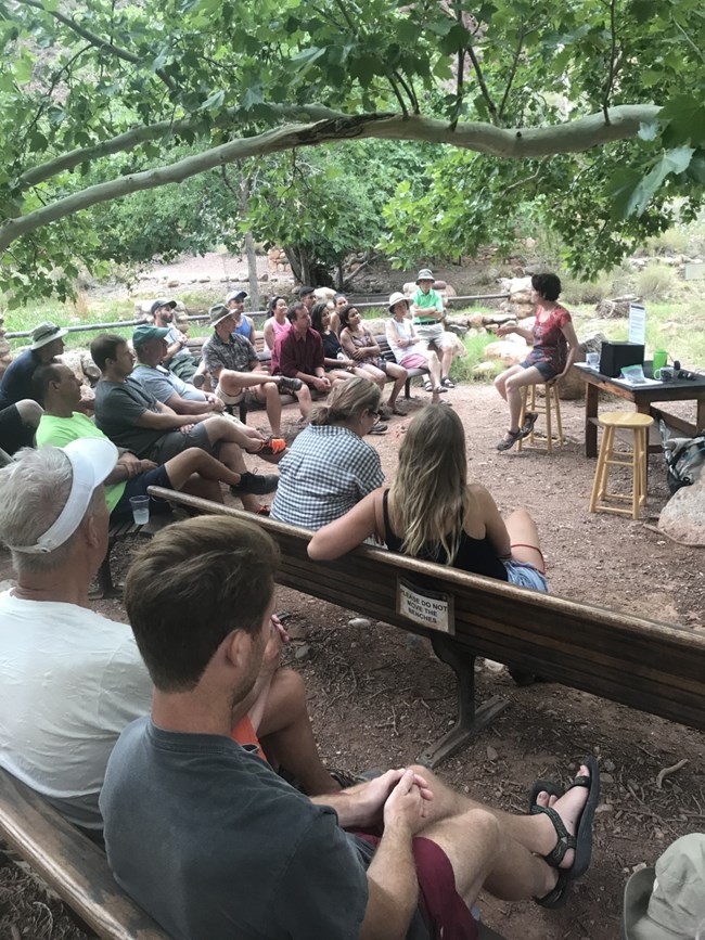 Twenty visitors sit around a sycamore tree while a woman speaks