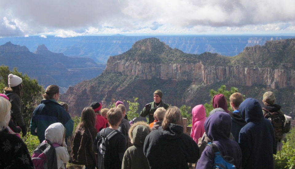 School group listening to a park ranger on the North Rim of Grand Canyon. a group of young student wearing jackets and hoodies are facing towards a park ranger that is giving a program. In the background, buttes and cliffs within a canyon landscape beneath a cloudy sky.