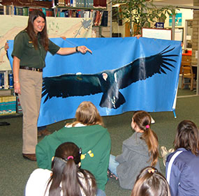 Students sitting on the floor listening to a female guest speaker who is standing to the left of a life-size banner of a California condor in flight. Students sitting on the floor listening to a female guest speaker who is standing to the left of a life-size banner of a California condor in flight.