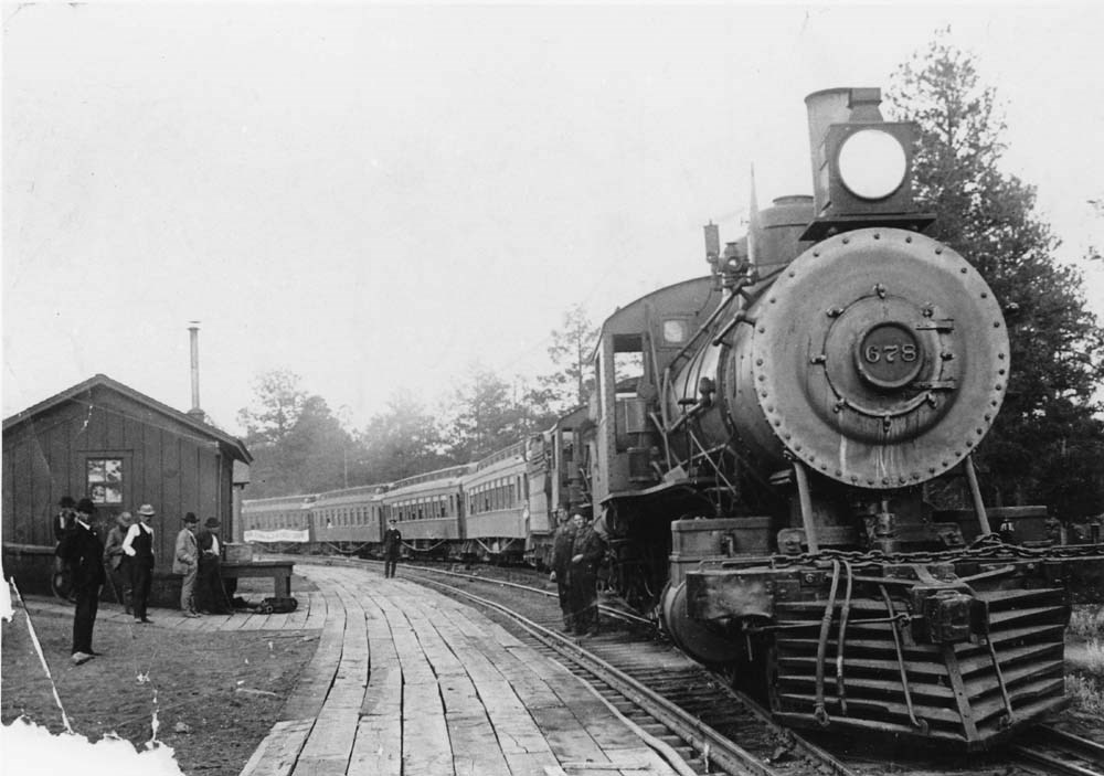 A steam engine pulls forward next to a wooden walkway with a small wood building. Pine trees are in the background. A steam engine pulls forward next to a wooden walkway with a small wood building. Pine trees are in the background.