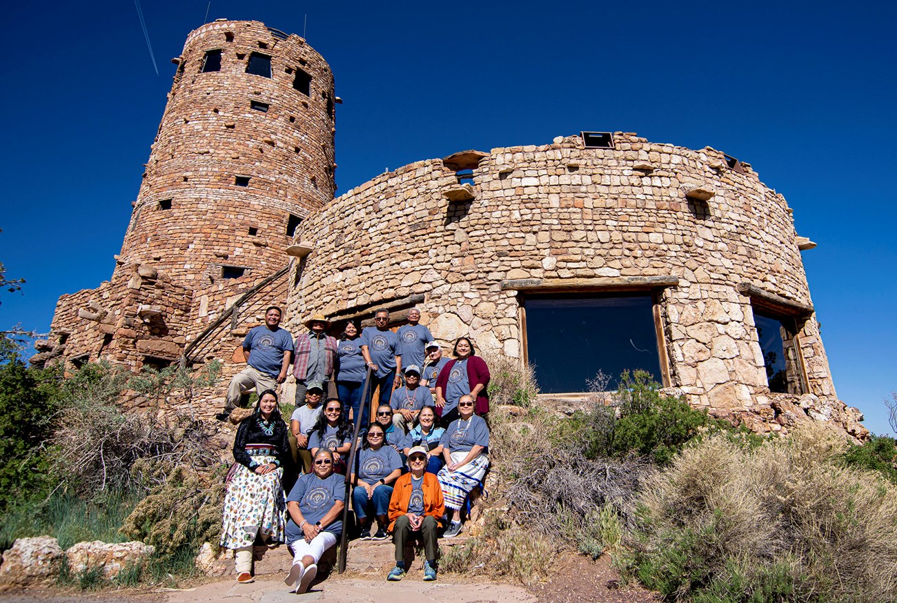 18 people arranged in three rows pose for a group photo on some steps leading to a stone building with a circular tower, in the style of ancestral Puebloan architecture.