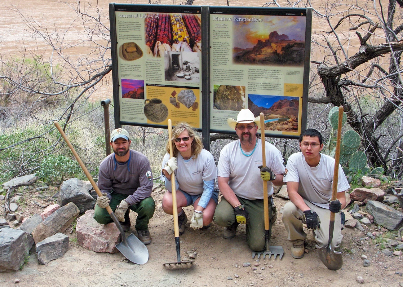 Bright Angel Pueblo Exhibit Installation Crew A group photo of 4 workers kneeling in front of two colorful outdoor exhibit panels interpreting an archaeology site. They are each posing while holding a shovel or a rake