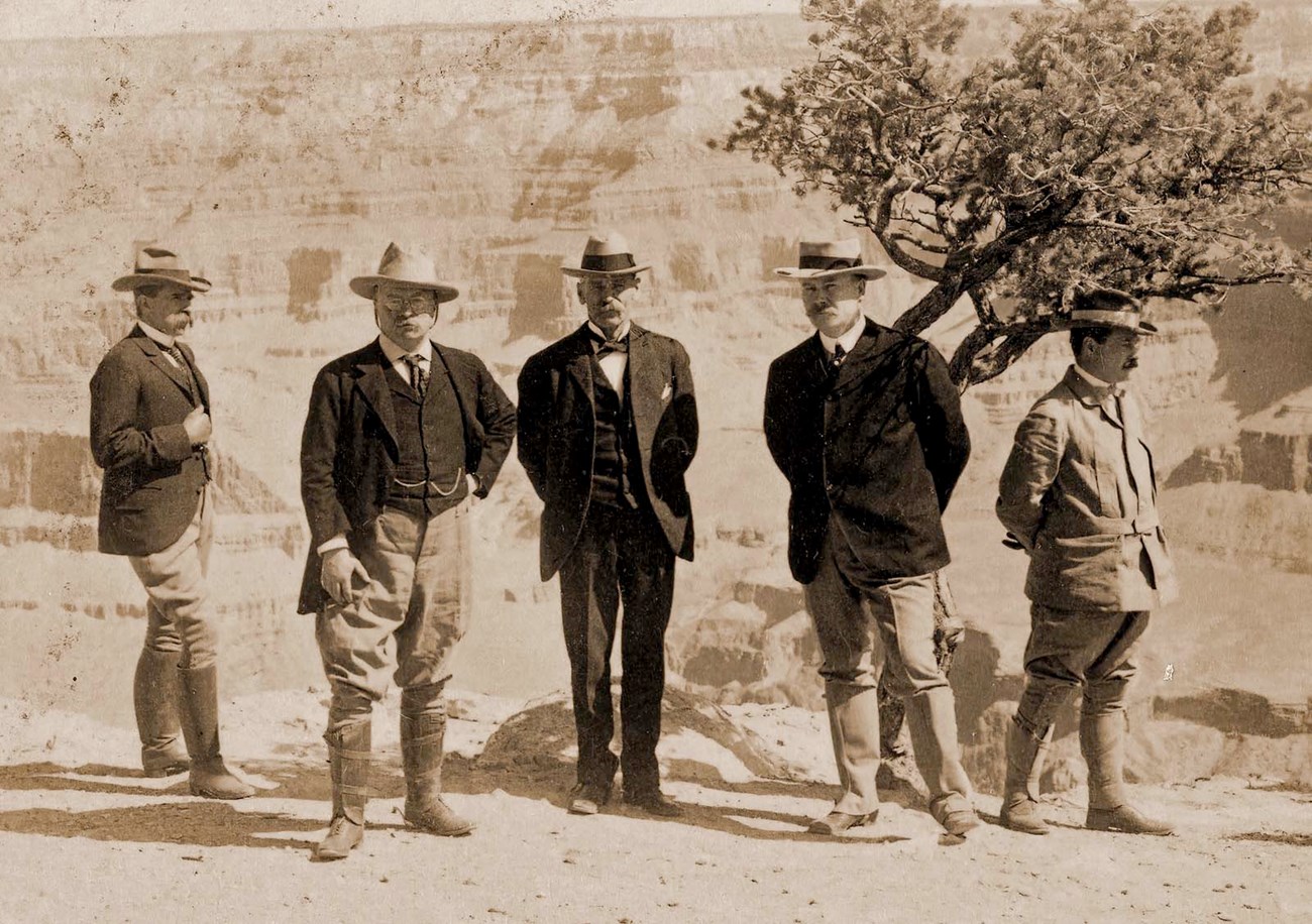 Historic sepia toned photo of 5 well dressed men wearing suits and standing in front of Grand Canyon. The second man from the left is President Theodore Roosevelt.