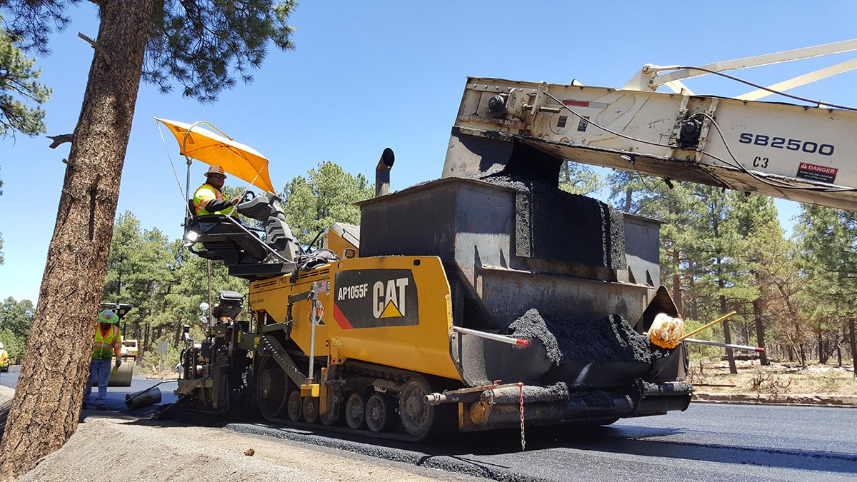 a large, yellow paving machine is spreading asphalt as it slowly moves down a highway. A Large white conveyor arm is transferring asphalt from another vehicle to the paving machine.