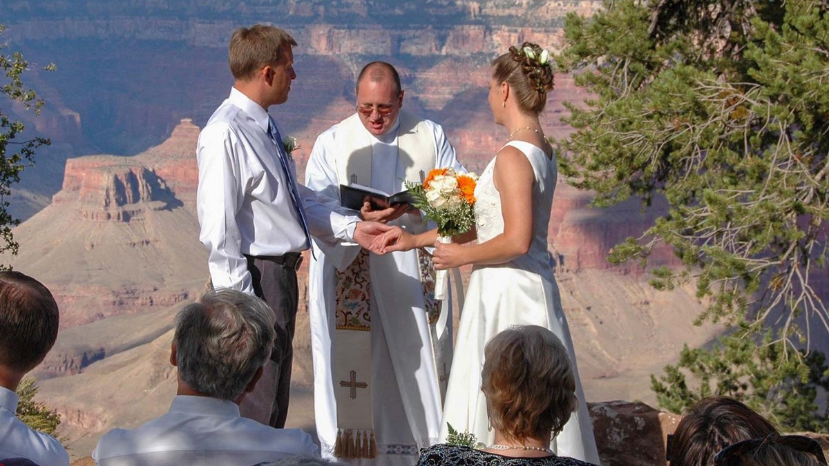 View past the heads people at a wedding ceremony with a bride and groom standing by a priest that is reading from a book. Colorful canyon buttes and cliffs form the background.