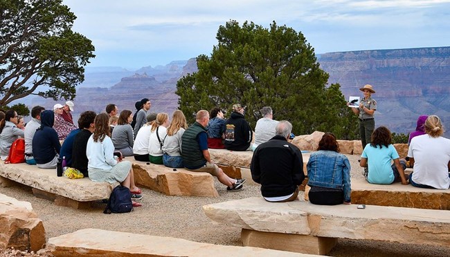 A park ranger shows holds a photo to show a crowd of visitors sitting around her during an evening presentation. Trees and a partial view of the Grand Canyon are visible in the background.