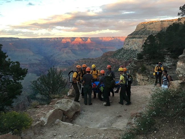 The sun sets in the background of the Grand Canyon, as a group of park rangers take a break carrying a litter up a trail during a search and rescue operation.