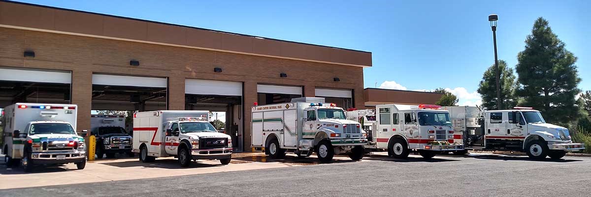 Six white emergency vehicles, fire trucks and ambulances and lined up in front of a large garage.