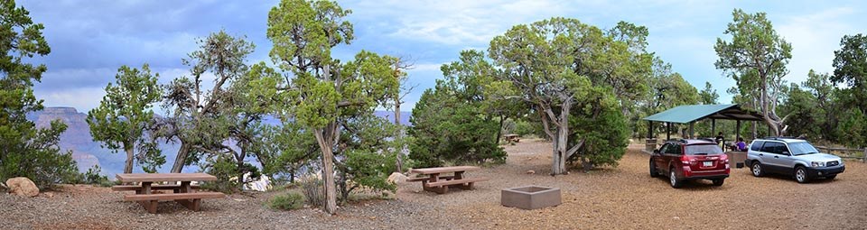 panoramic photo of a special event site on the edge of a canyon. There are several picnic tables under pinyon and juniper trees, and a picnic shelter with 2 cars parked nearby.
