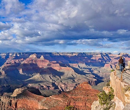 Several sightseers behind guardrails at a scenic overlook above a mile deep canyon filled with colorful peaks and cliffs.