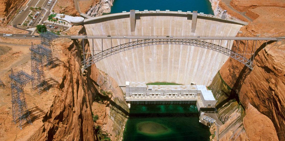 Aerial view looking down on a large concrete dam between orange colored rock walls. a suspension bridge is in the foreground, with cars driving across.