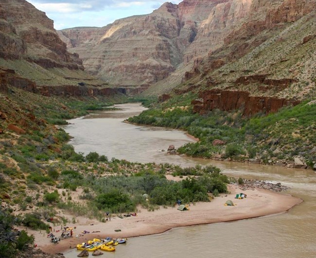Beneath canyon walls, a river makes a bend around to a large sandbar where boaters are camping. 6 rafts are tied to the shore an several tents are set up on the sand.