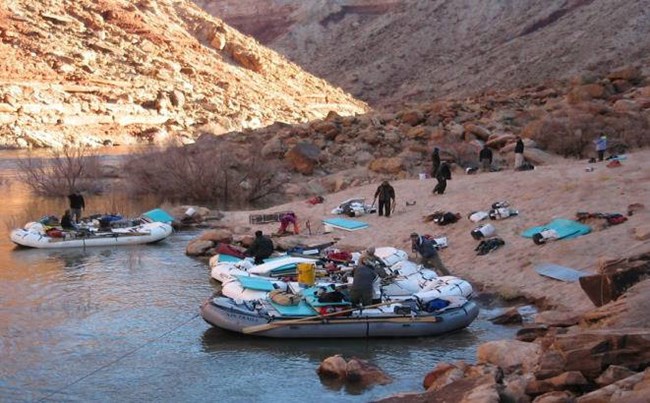 a river camp with 5 grey rafts tied to the shoreline of a muddy river. beyond the boats a flat area where kitchen tables have been set up.